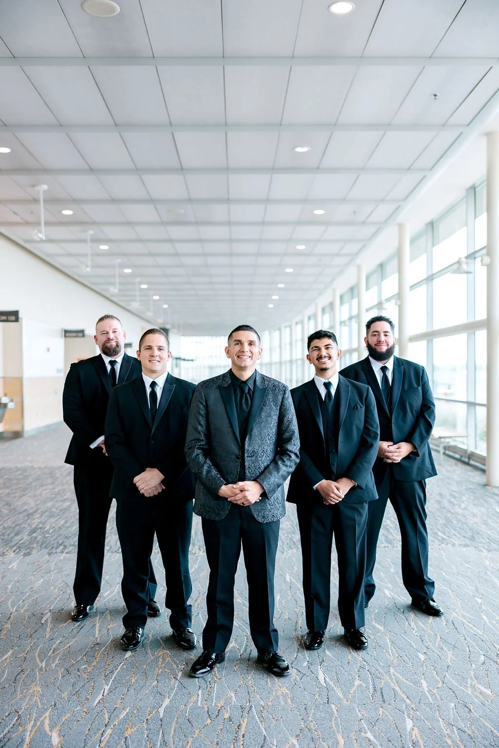 A group of five men dressed in formal black suits standing indoors in a well-lit, modern hallway with large windows and a patterned carpet.