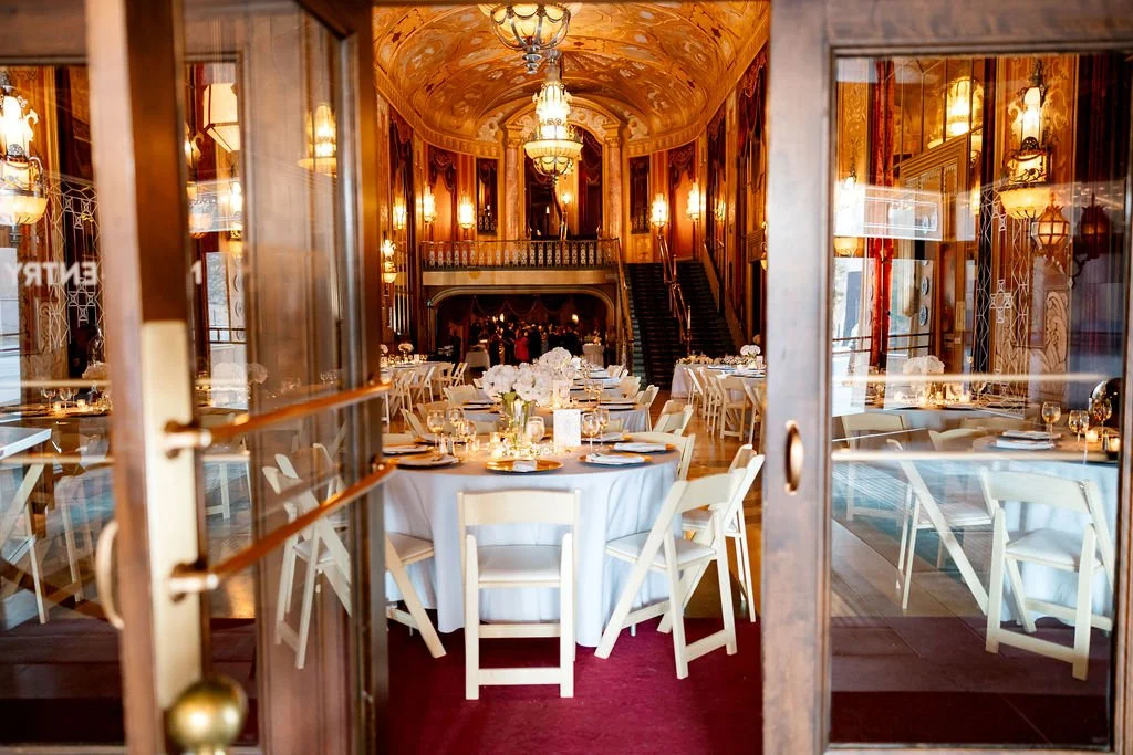 Elegant banquet hall with high ornate ceilings, chandeliers, and decorated walls, seen through glass doors. Banquet tables are set with white tablecloths, floral centerpieces, and place settings, ready for a formal event.