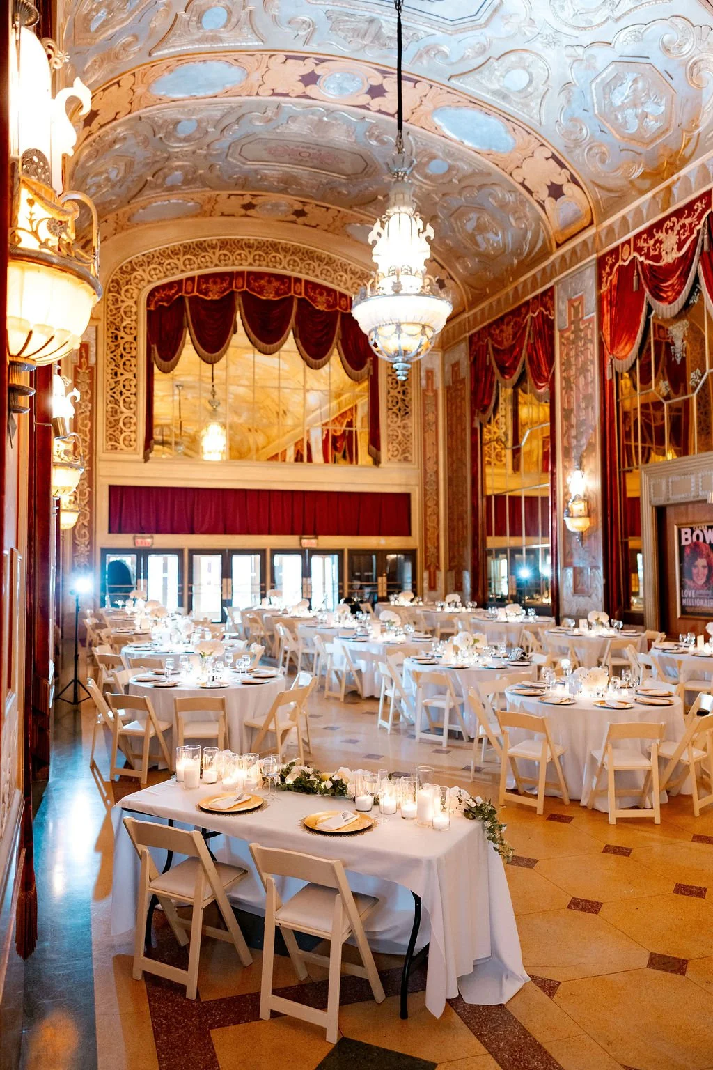Elegant ballroom with high, ornate ceiling, chandeliers, and red velvet curtains. Round tables with white tablecloths and floral centerpieces are set for a formal event, with candles and dinnerware. A long head table at the front is decorated with ca