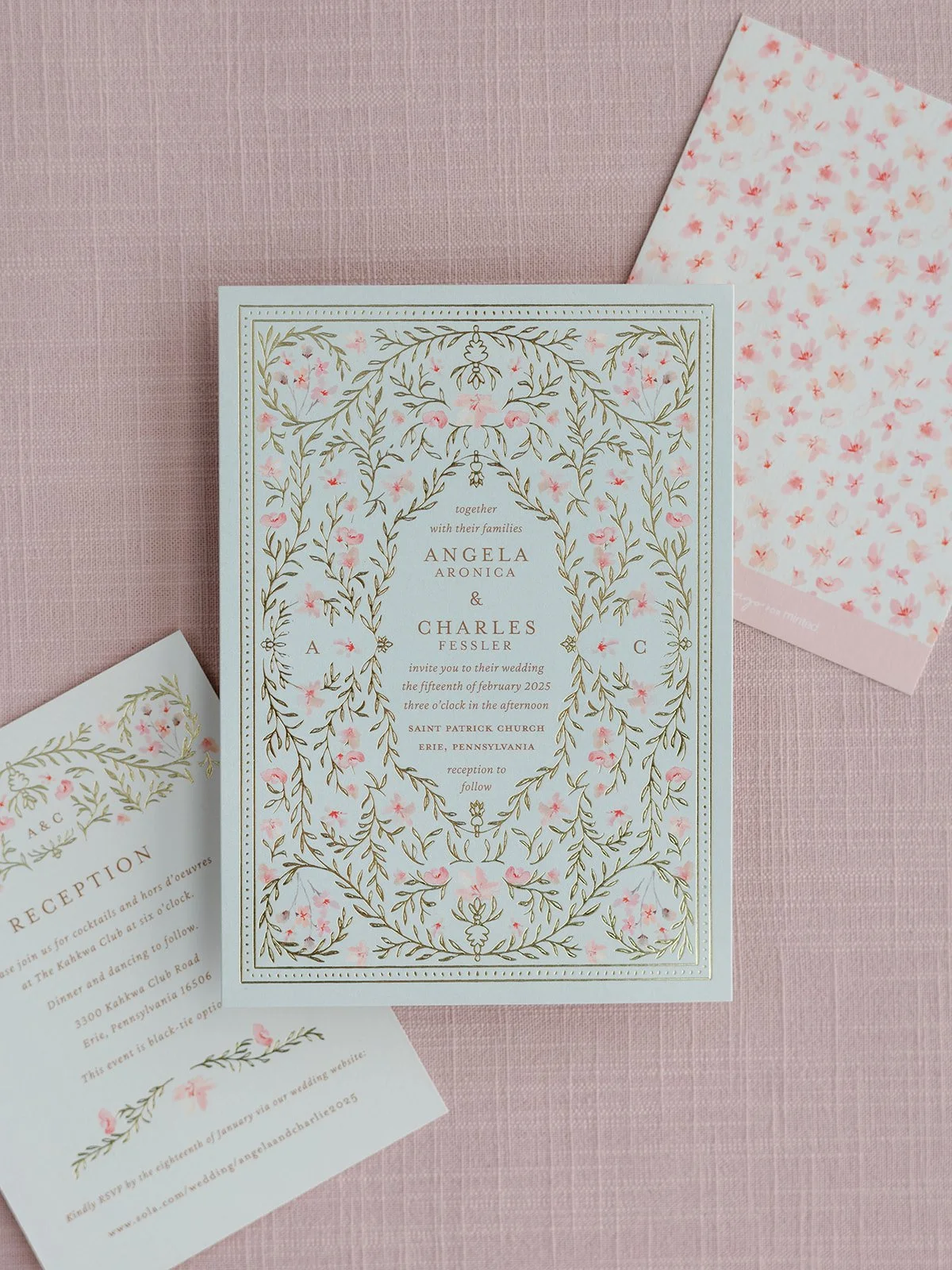 Wedding invitation with floral design, gold accents, and pink flowers, laying on pink fabric, with matching RSVP card and floral envelope.