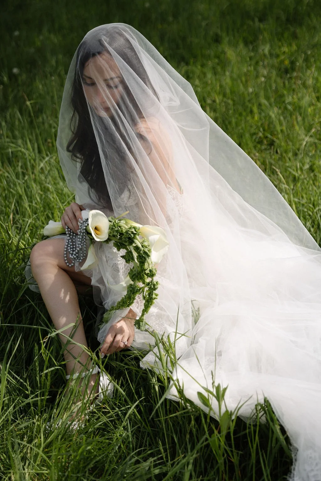 Woman in a wedding dress and veil holding a bouquet of white calla lilies and greenery, sitting on grass in a field.