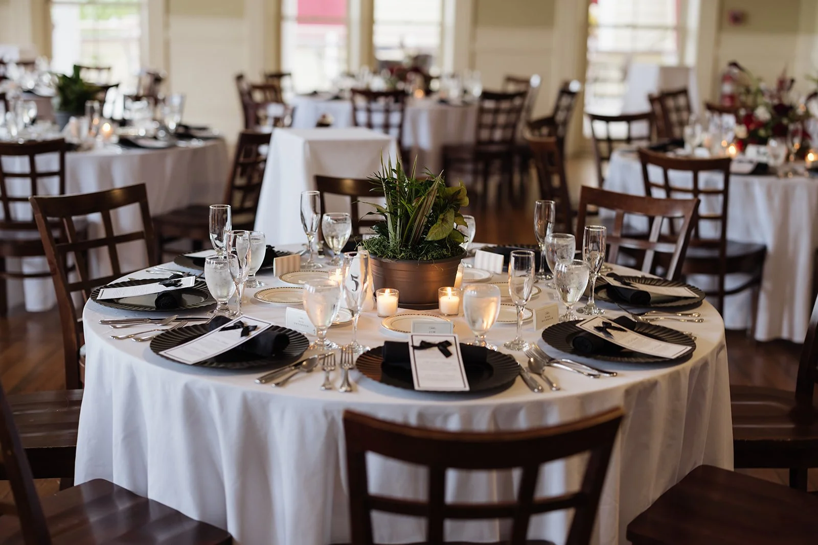 Elegant banquet table setup with black plates, silverware, glasses, black napkins, a green centerpiece, and lit candles in a well-lit room with multiple similar tables.