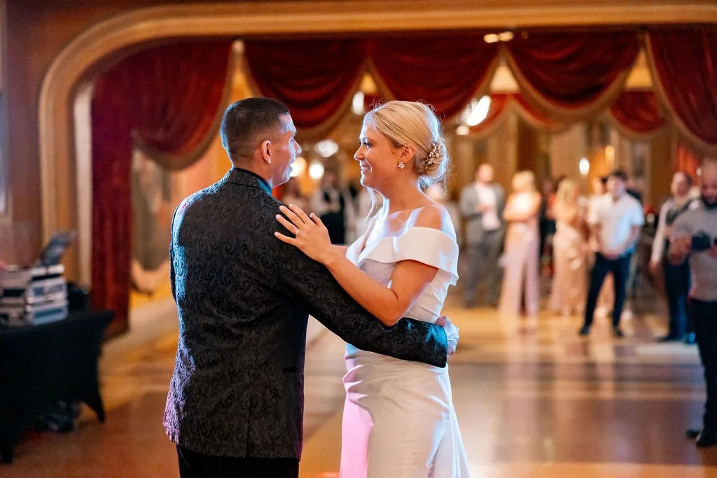 A newly married couple sharing their first dance at a wedding reception in a decorated ballroom with red curtains and guests in the background.