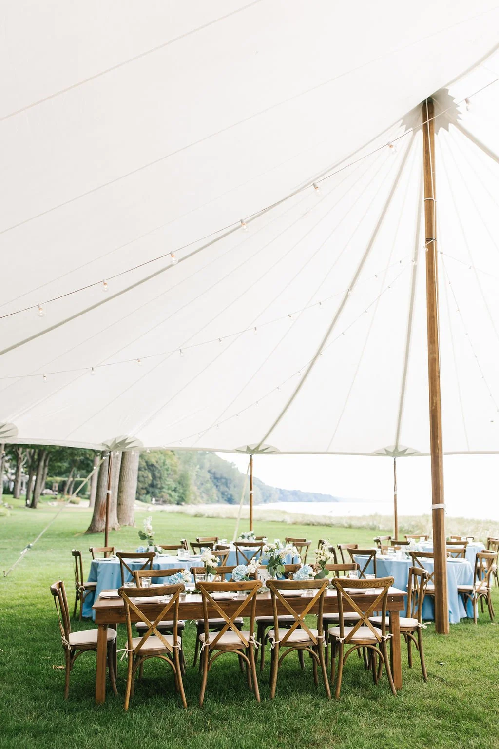 Outdoor event setup with wooden tables and chairs under a large white canopy tent, decorated with flower centerpieces on the tables, on a grassy area near trees by a lake.