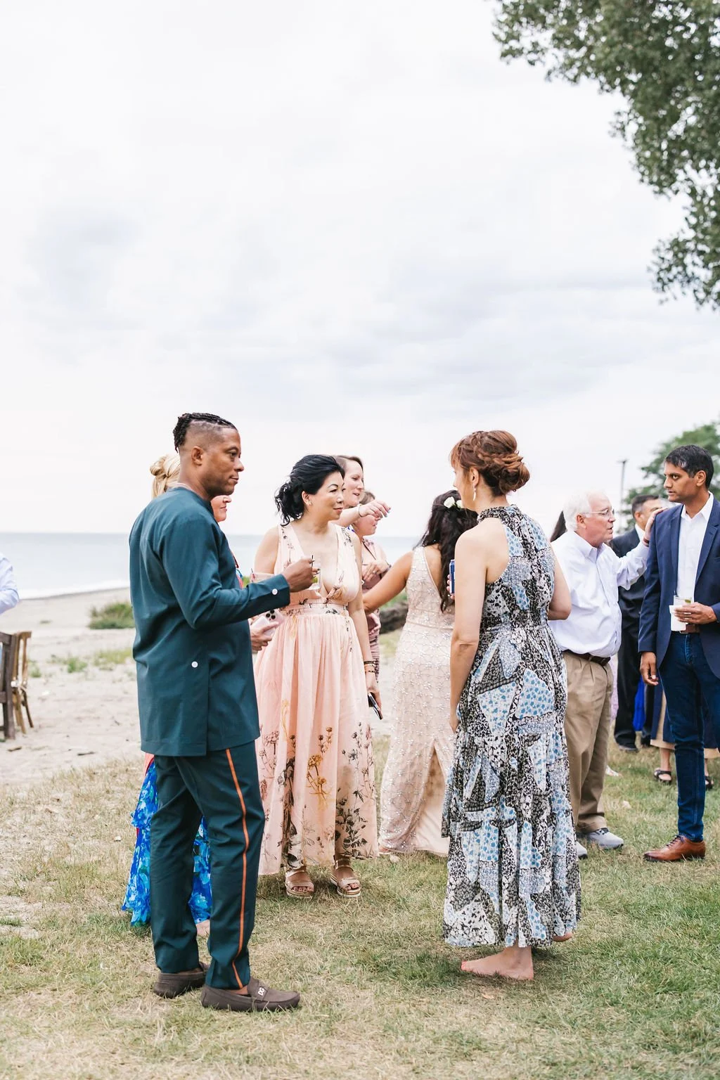 A group of people dressed in formal and semi-formal attire socializing outdoors near a beach, with some holding drinks, on a cloudy day.