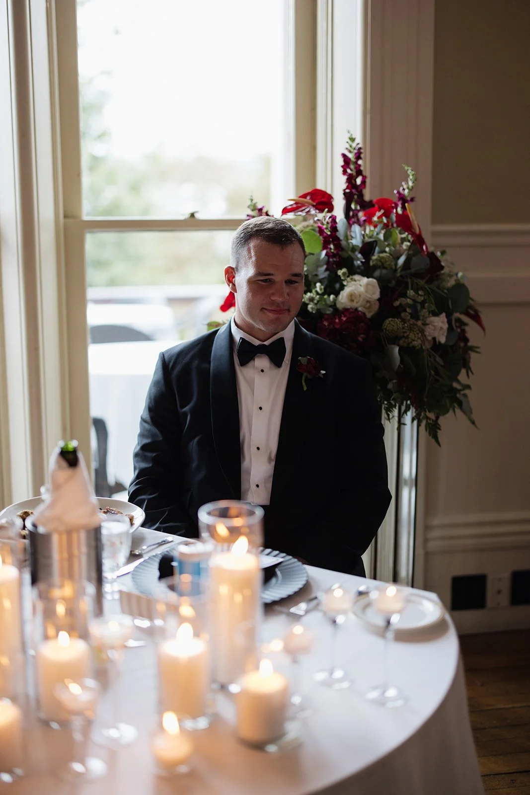 A groom in a black tuxedo and bow tie sitting at a decorated wedding reception table with lit candles. There is a large floral arrangement called a centerpiece, and a window showing outdoor scenery in the background.