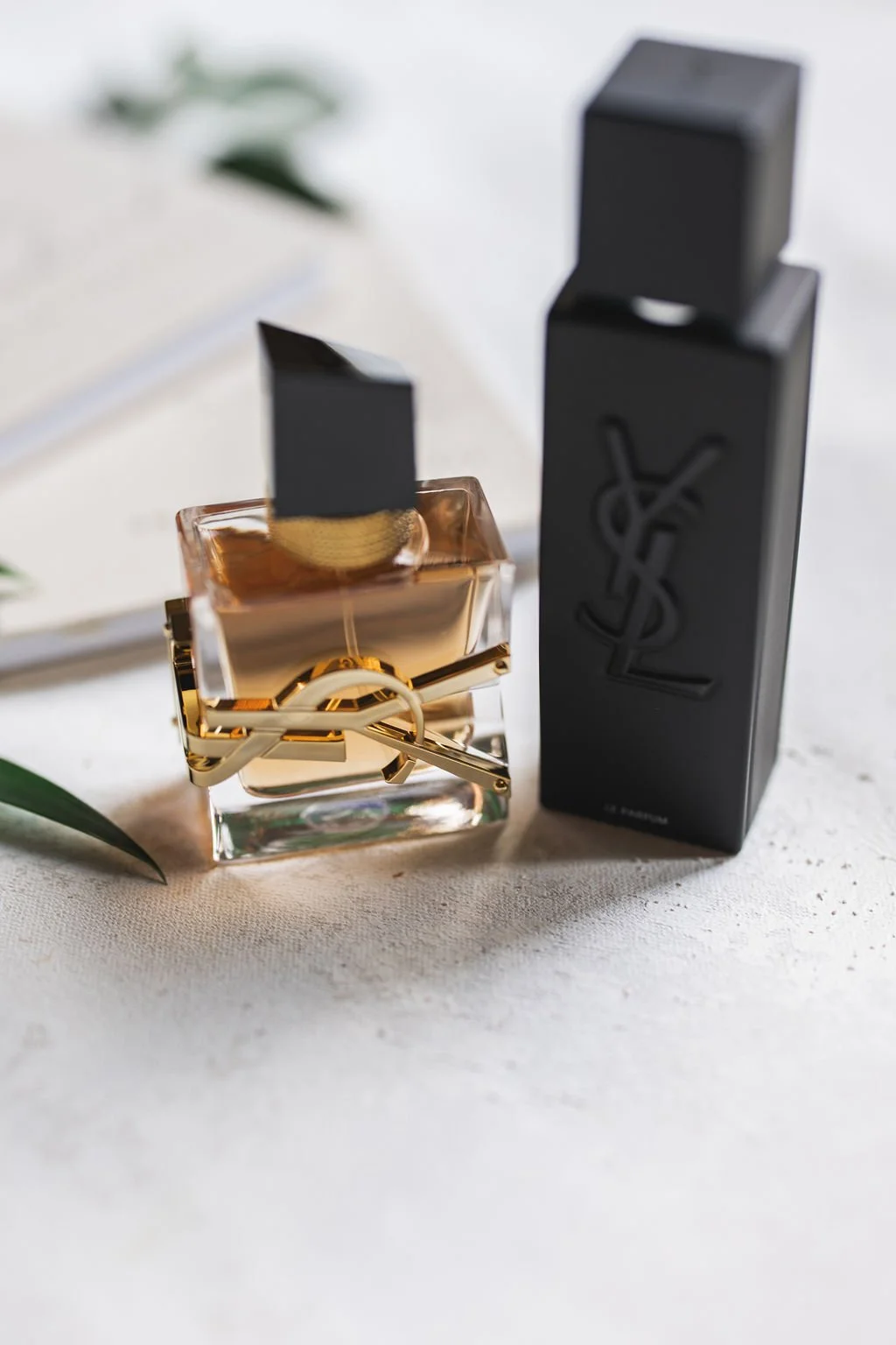 A bottle of Yves Saint Laurent Black Opium perfume and its black packaging box on a white surface with a plant leaf in the background.
