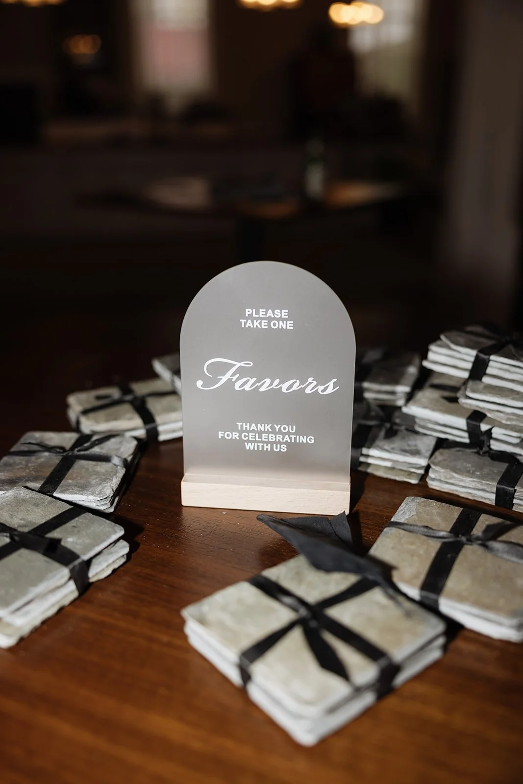 A table with multiple stacks of small black-and-white napkins tied with black ribbon, and a sign that says "Please Take One. Favors. Thank you for celebrating with us."
