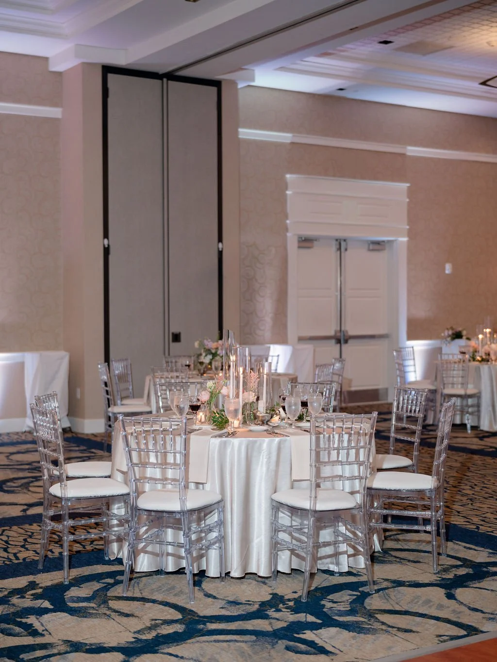 Wedding reception setup with round tables covered in white tablecloths, set with glassware, plates, silverware, floral centerpieces, and candles, in an elegant banquet hall with patterned carpet and beige walls.