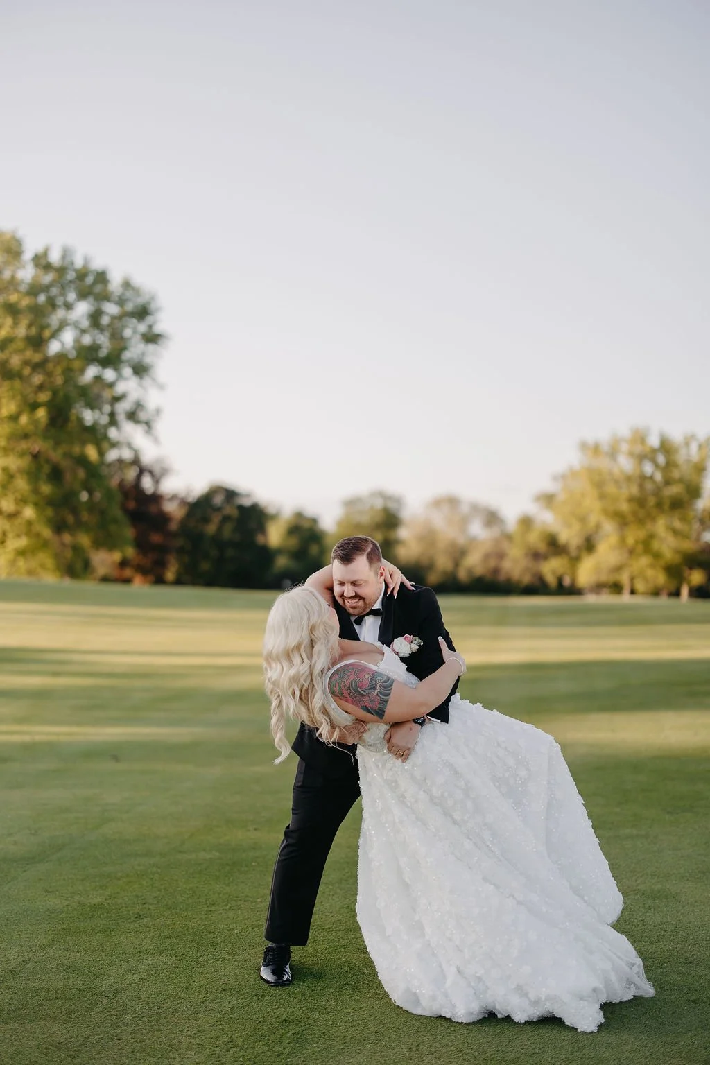 A newlywed couple dancing outdoors on a grassy field during sunset, with trees in the background.