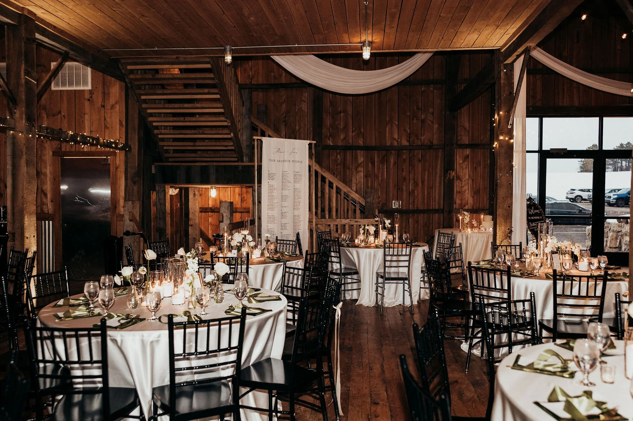 Wedding reception setup with round tables covered in white tablecloths, decorated with candles, floral arrangements, and place settings, in a rustic wooden venue with high ceilings, wooden walls, string lights, and windows showing a parking lot outsi