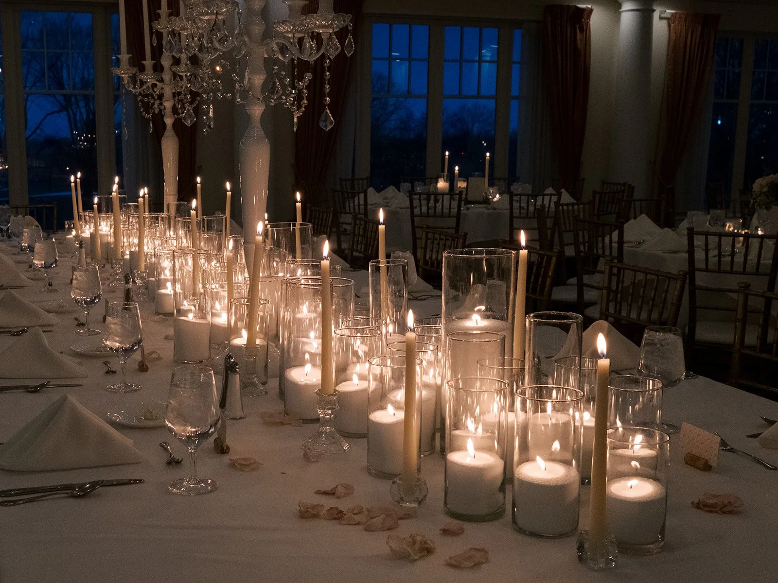 A dimly lit banquet table decorated with numerous tall candles in glass holders, creating a warm, romantic ambiance. The table is set with white napkins, glassware, and scattered rose petals. In the background, large windows reveal a dark evening sky