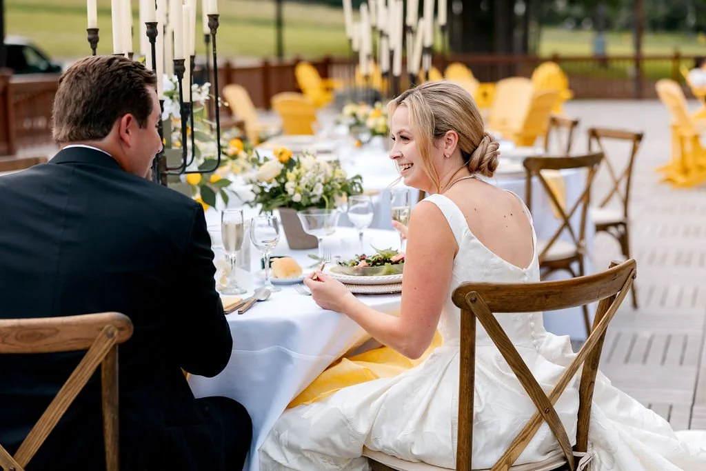 A bride and groom dining at an outdoor wedding reception table decorated with flowers, candles, and tableware.