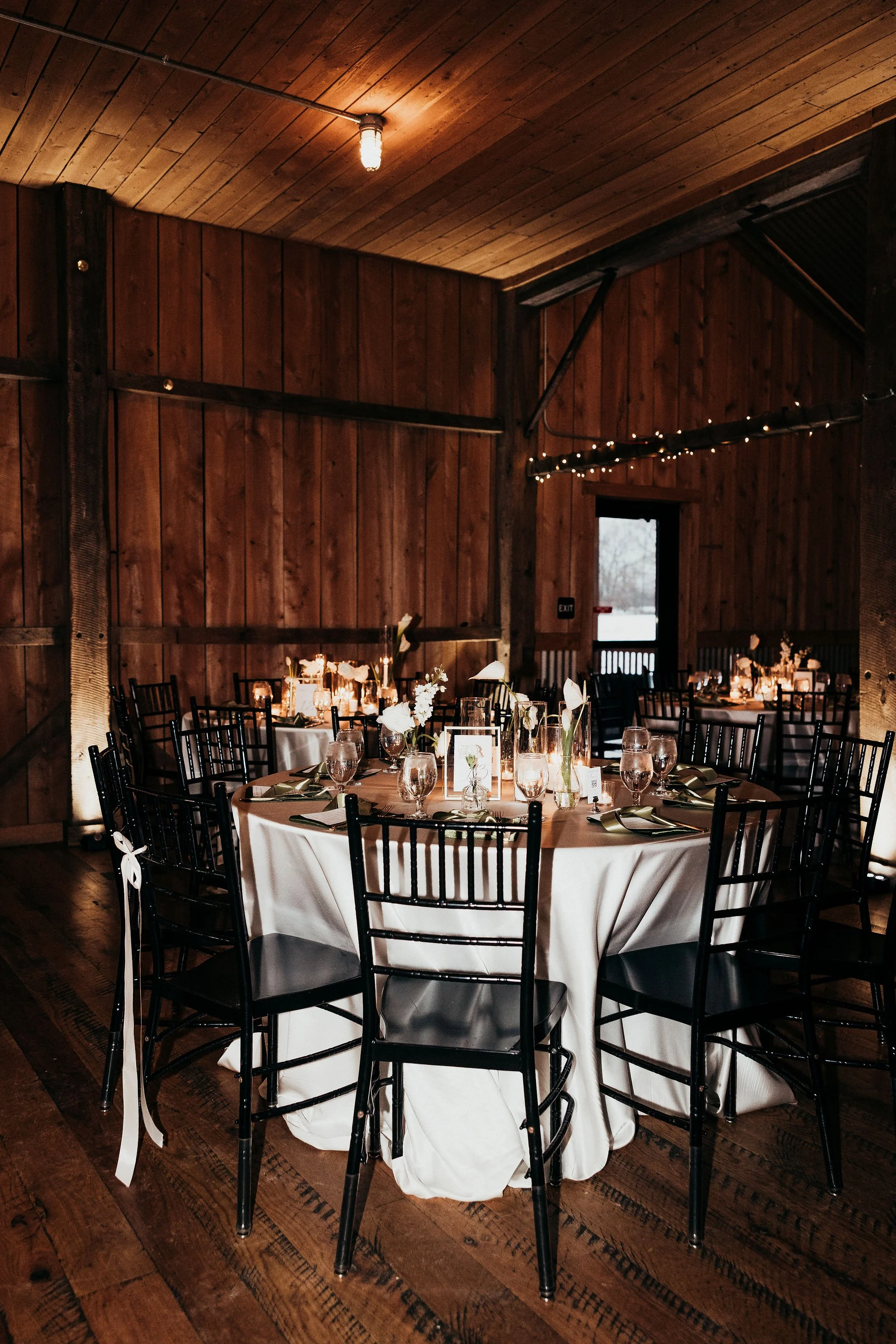 A round table covered with a white tablecloth, decorated with candles and floral centerpieces, set for a formal event in a wood-paneled banquet hall with black chairs and string lights.