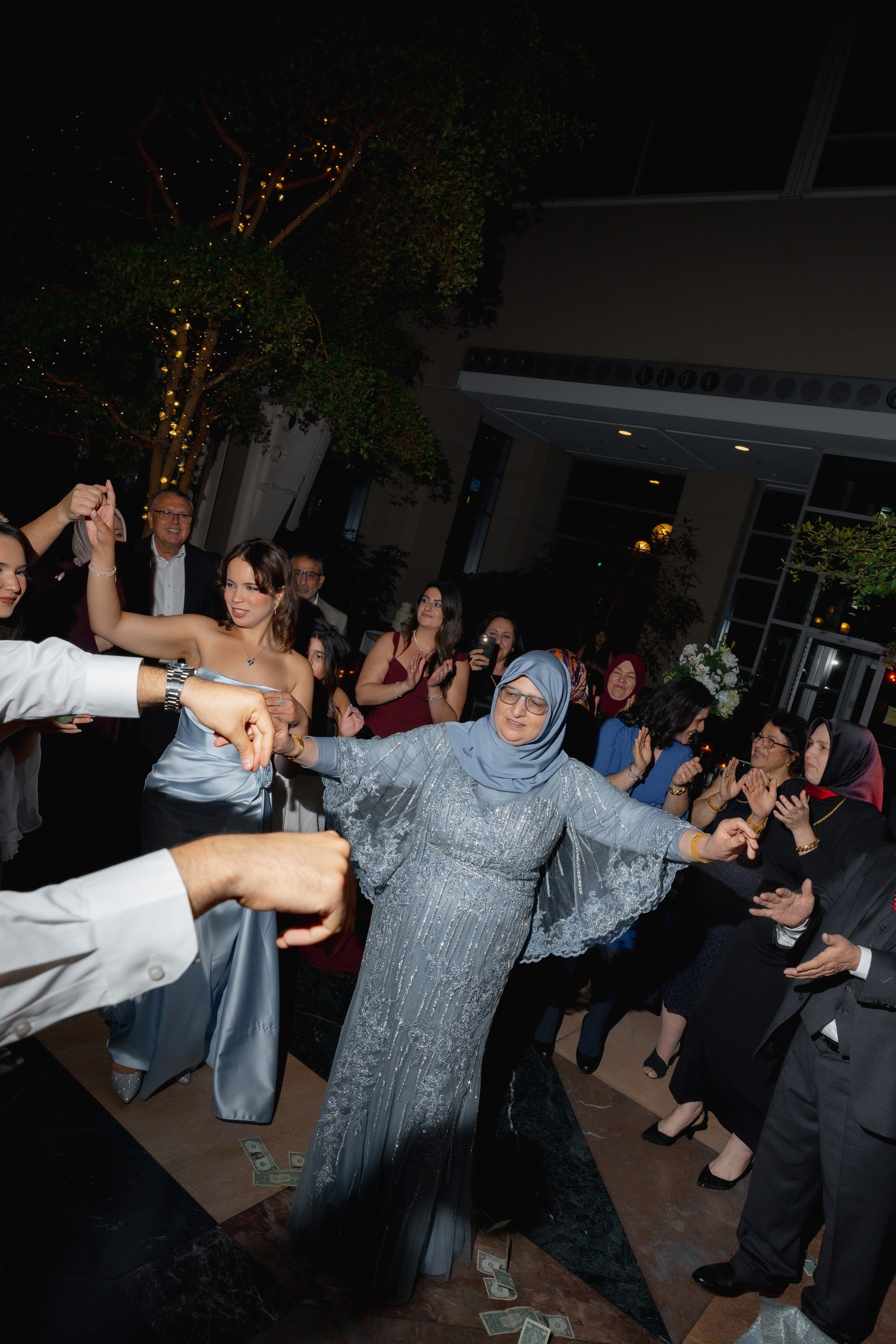 A group of people dancing at an indoor celebration event, with some holding hands in a circle, and dollar bills scattered on the floor.
