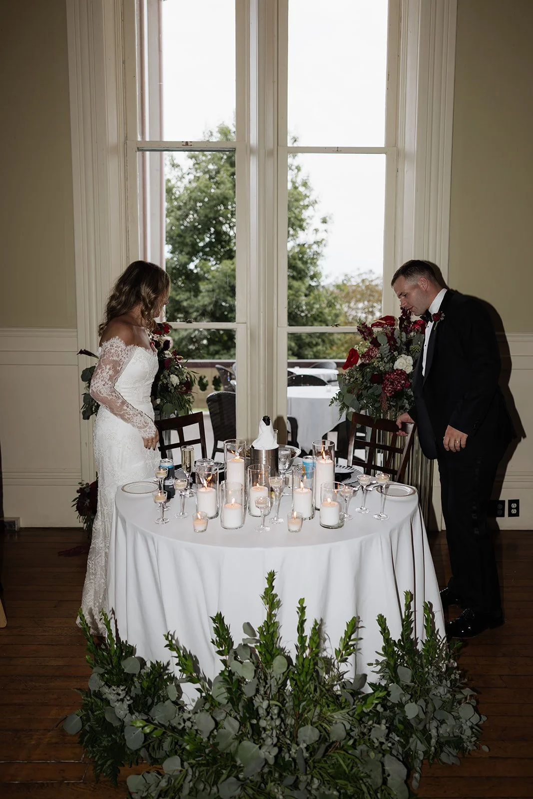 A bride and groom standing on opposite sides of a table with candles and floral arrangements during a wedding ceremony, indoors in front of a large window with trees outside.
