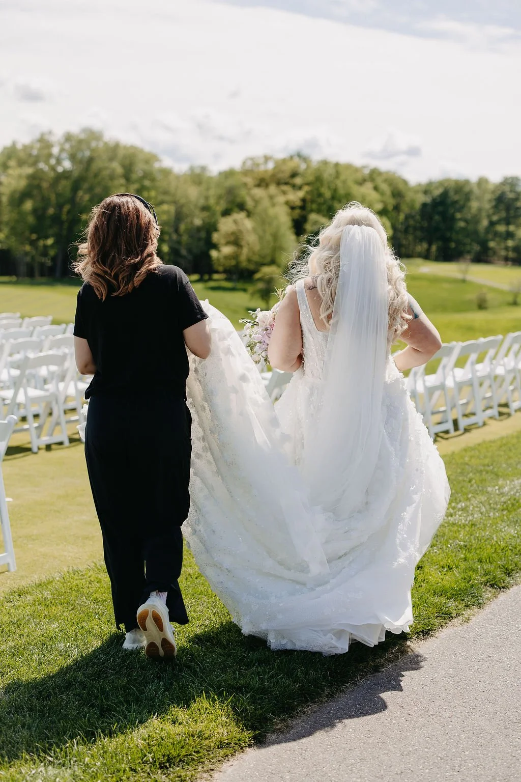 Two women, one in a white wedding dress with a long veil, the other in black, walk side by side outdoors on green grass with white chairs and a scenic background.