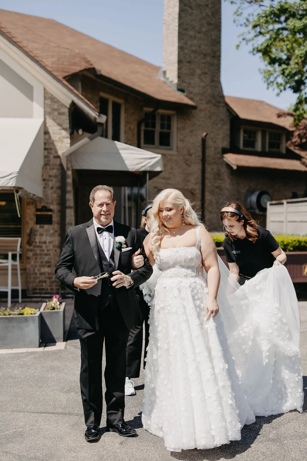 Bride in a white wedding gown walking with a man in a tuxedo, accompanied by a woman adjusting her dress, outdoors in front of a brick house on a sunny day.