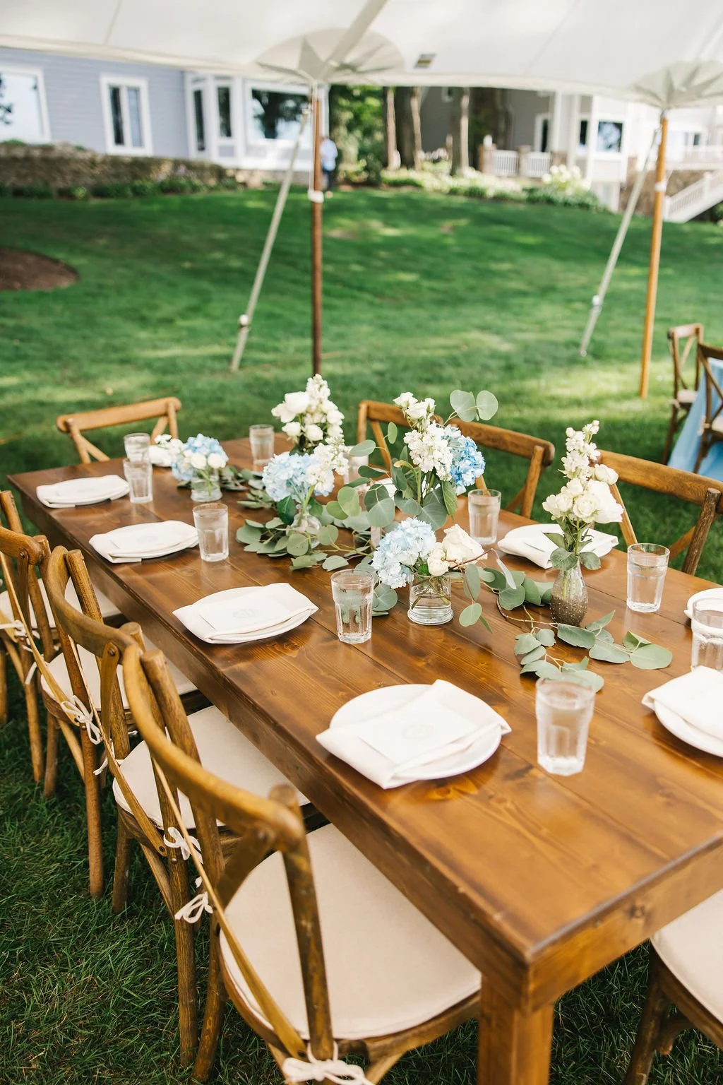 Outdoor dining table decorated with white and blue floral arrangements, set with white napkins, plates, and glasses, surrounded by wooden chairs on a grassy lawn under a tent.