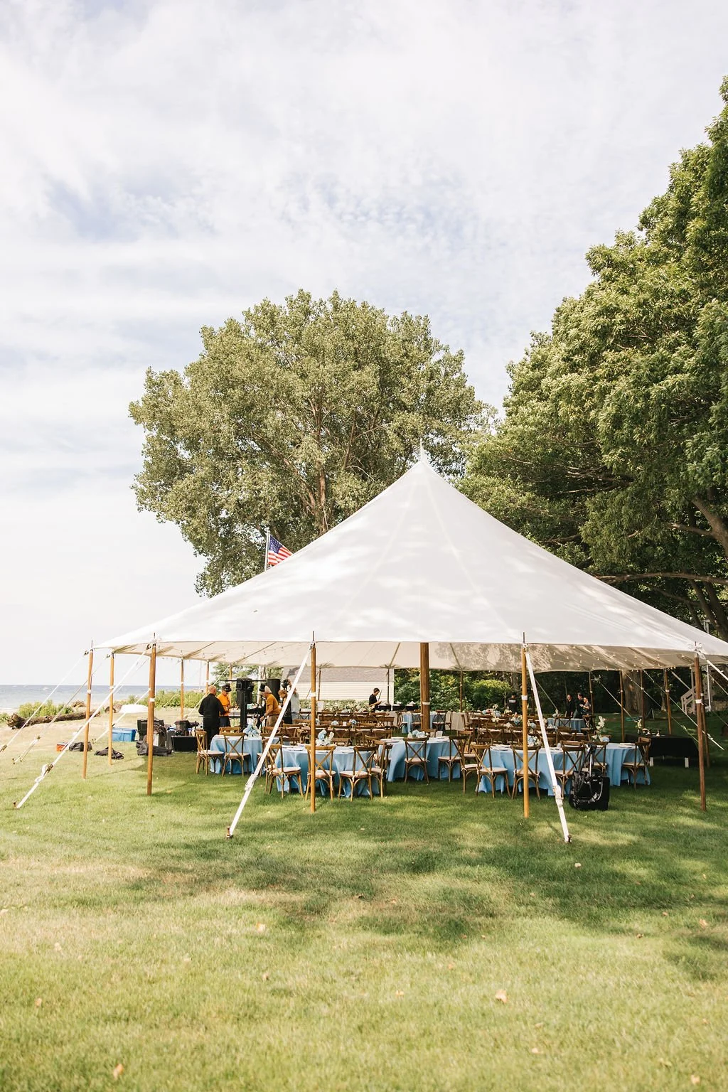 Outdoor event setup with a large white tent, tables with blue tablecloths, wooden chairs, and an American flag, surrounded by green trees on a grassy lawn under a partly cloudy sky.