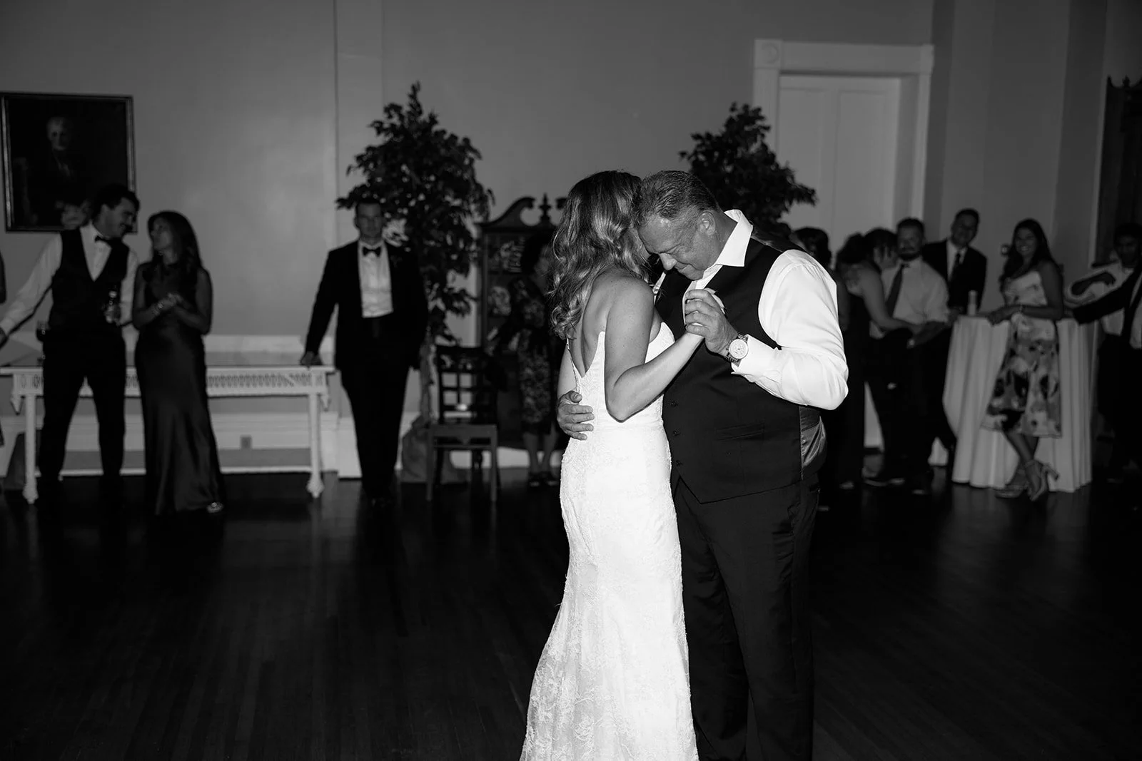 Black and white photo of a bride and groom dancing closely at their wedding reception, surrounded by well-dressed guests.