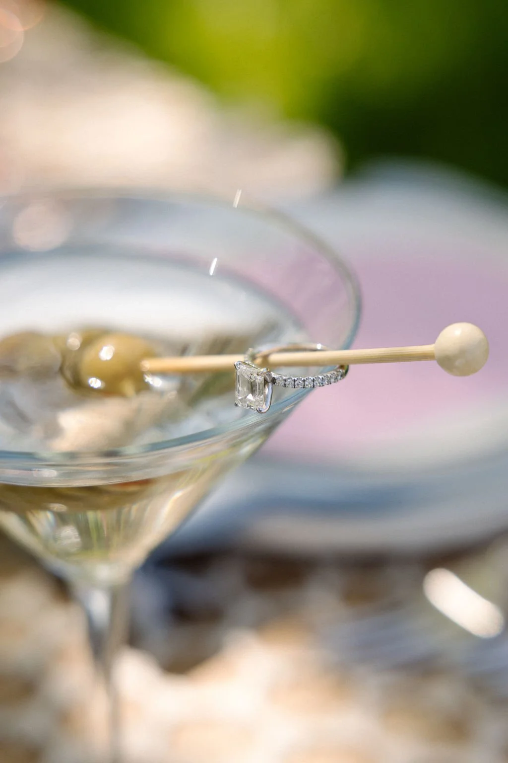 A martini glass filled with a clear beverage, garnished with an olive on a toothpick, and a diamond ring draped over the glass rim, with a blurred background.