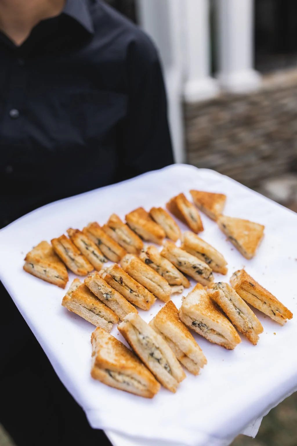 A person in a black shirt holding a white tray with assorted sandwich wedges, possibly a platter for serving.
