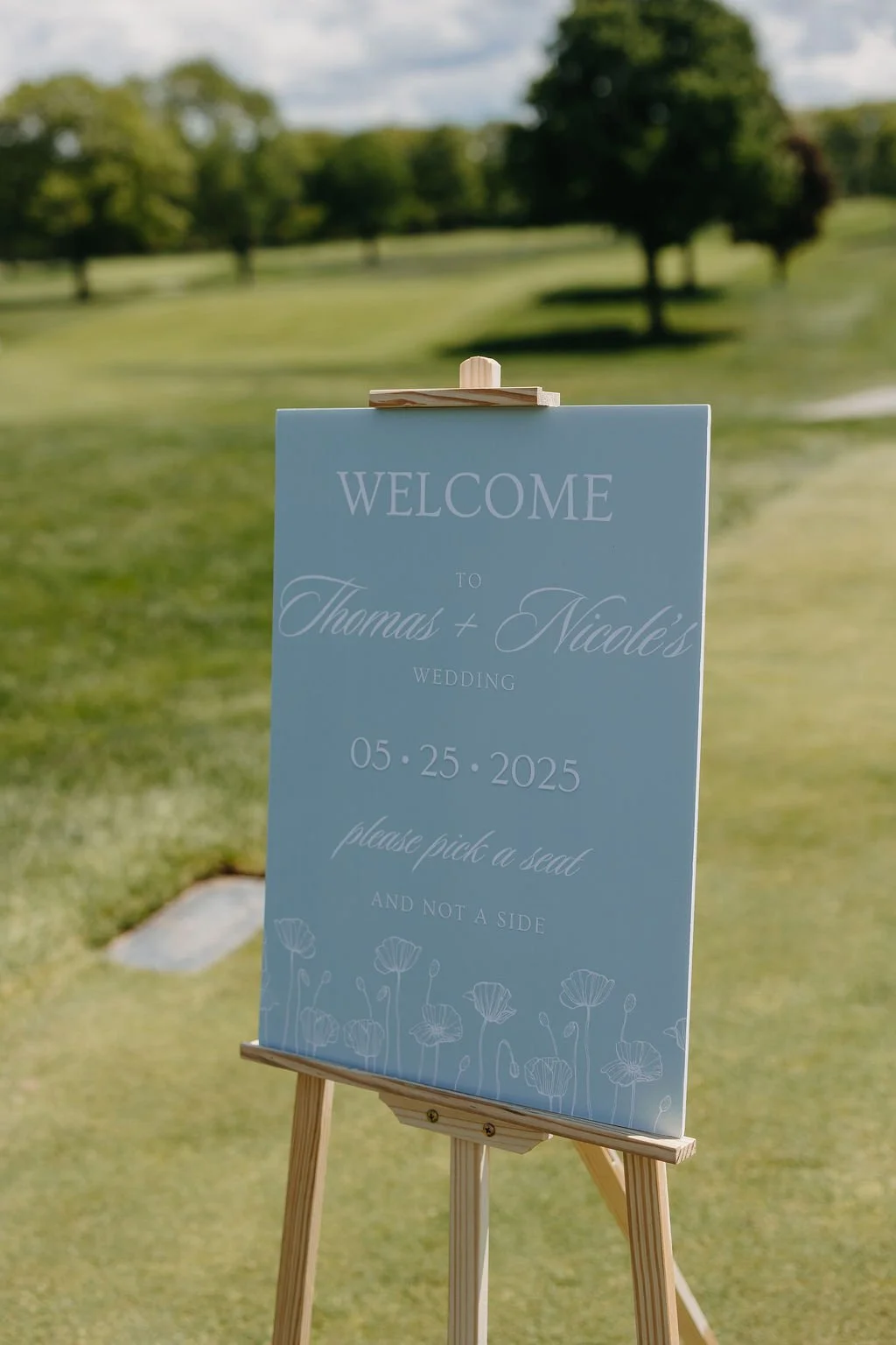 Wedding welcome sign on an easel outdoors with trees and grass in the background. The sign says "Welcome to Thomas & Nicole's Wedding 05.25.2025" and includes a request to "please pick a seat and not a side." The sign features light pastel colors and