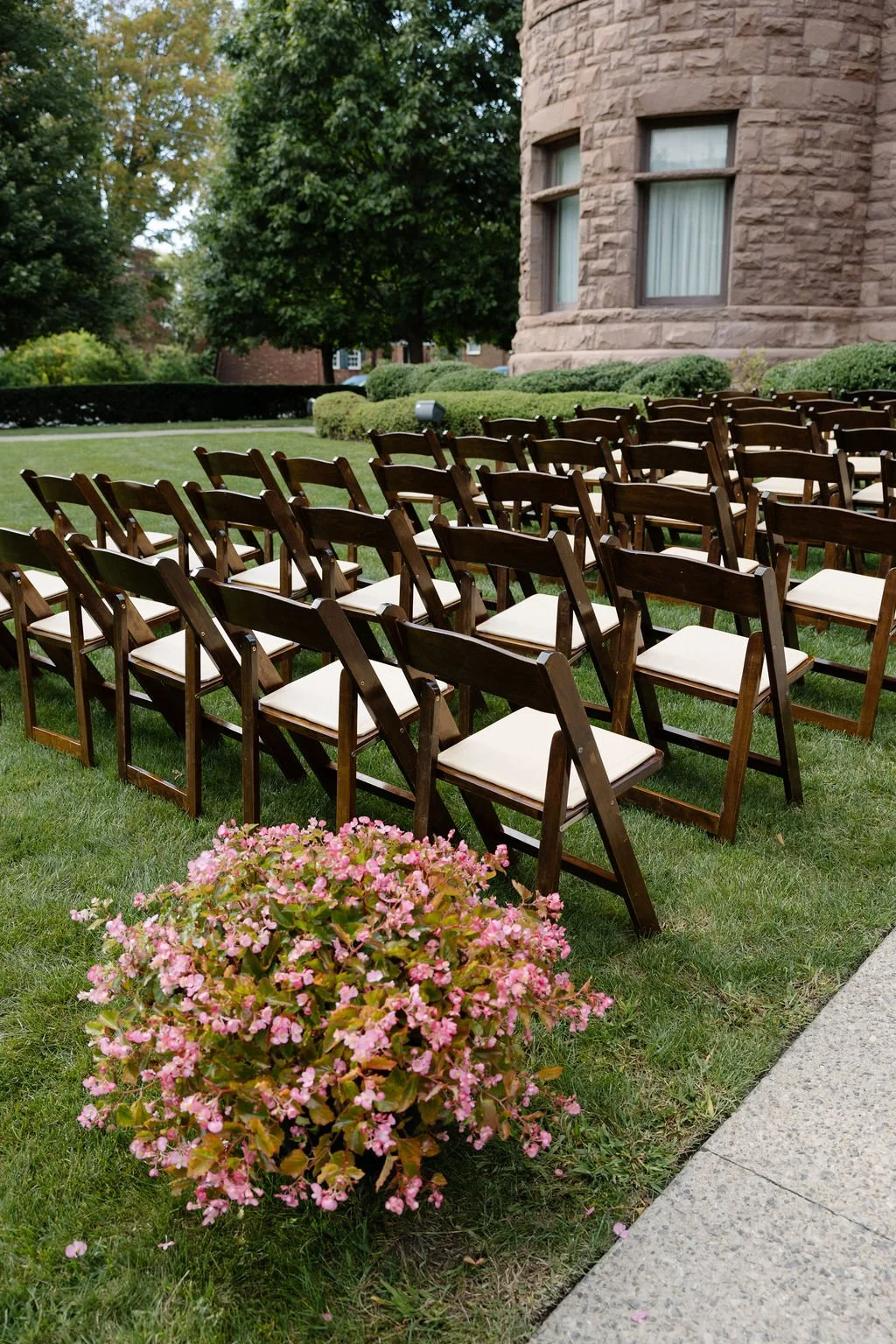 Outdoor setup with several rows of wooden chairs with white cushions on a grassy area, near a large brick building with multi-pane windows, surrounded by trees and shrubbery, possibly prepared for a ceremony or event.