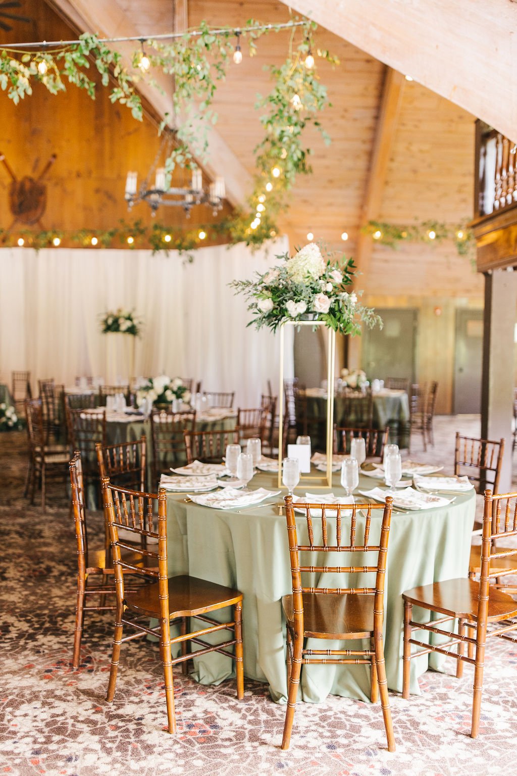 Elegant indoor wedding reception with round tables decorated with green tablecloths, white floral centerpieces, and wooden chairs, under string lights and greenery decorations in a rustic space.