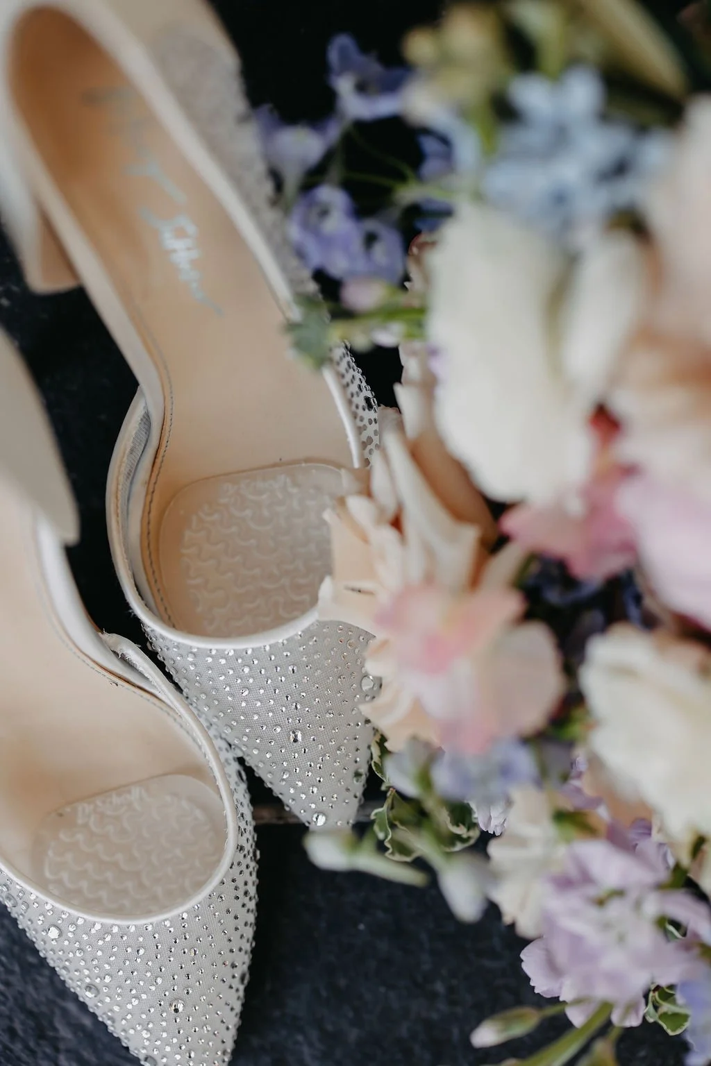 A pair of beige high-heeled shoes decorated with small rhinestones, surrounded by pink, white, and purple flowers on a dark surface.