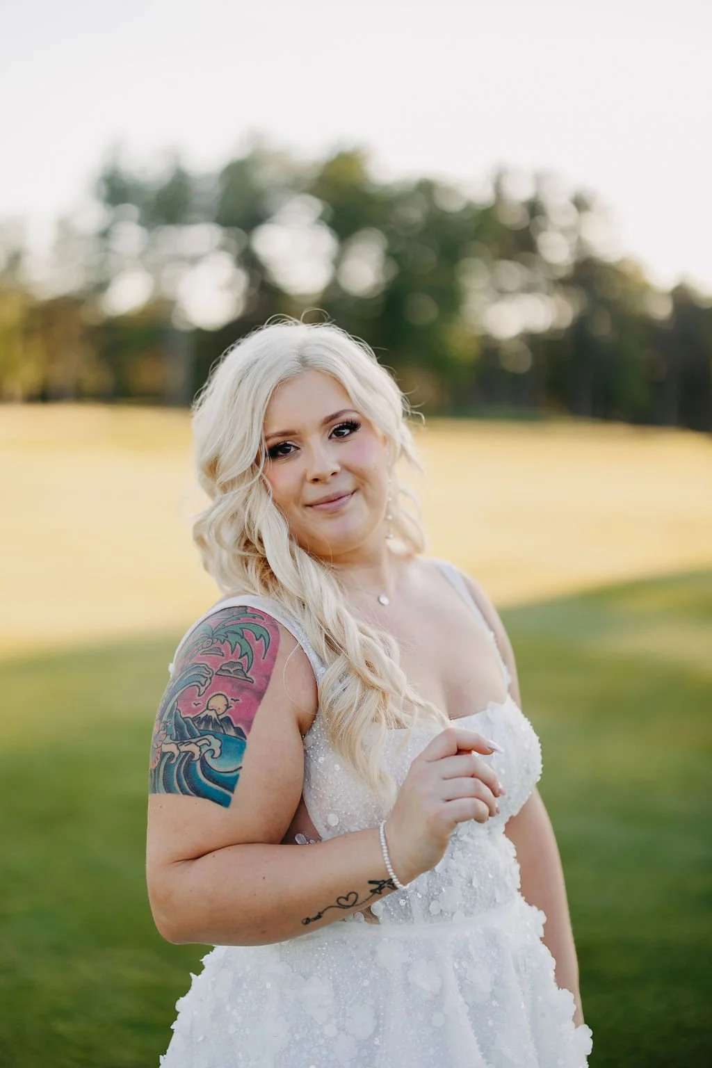 A smiling woman with long blonde curly hair wearing a white dress outdoors with a blurred green and yellow background.