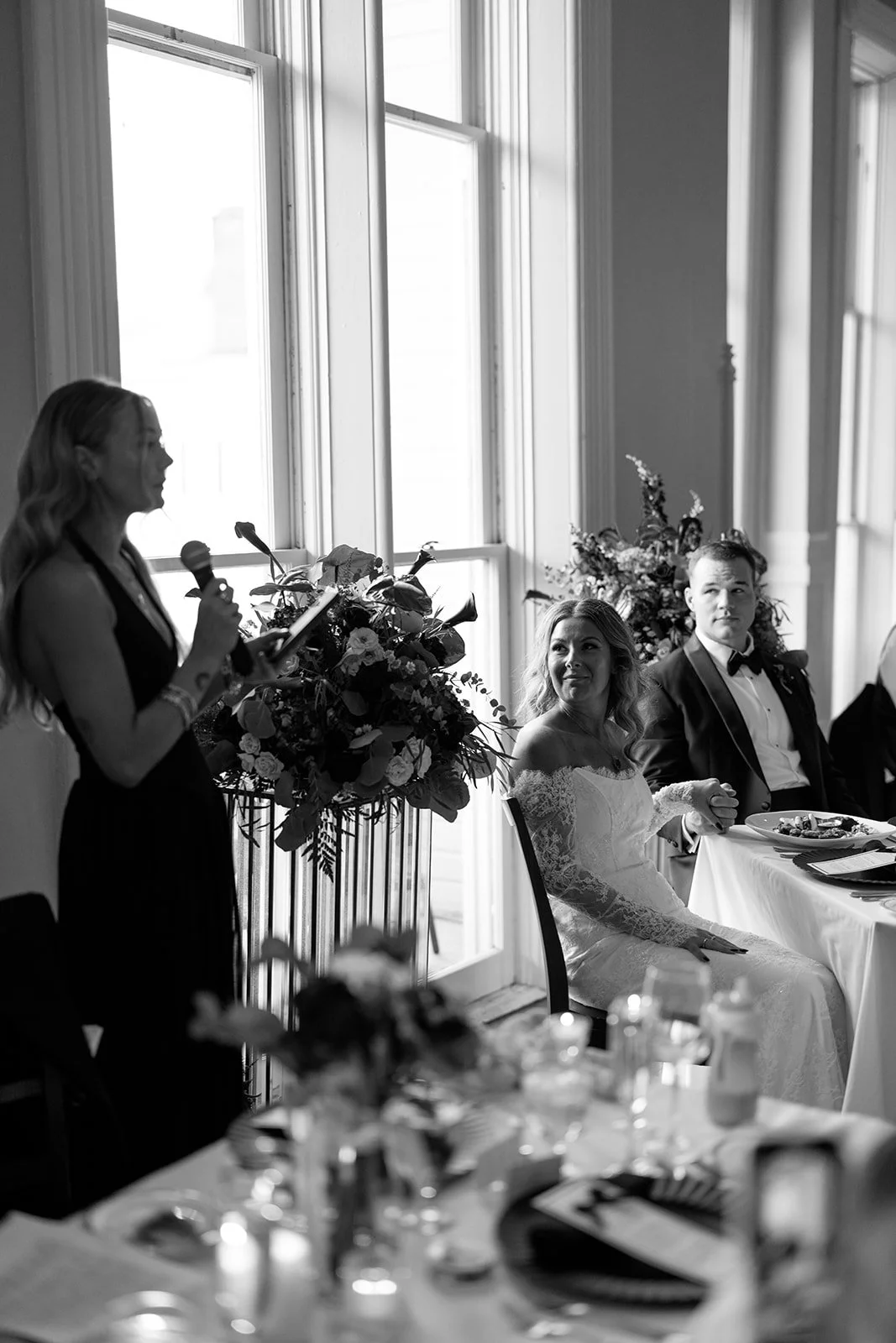 A woman giving a speech at a wedding reception, with a bride and groom seated at a table listening.