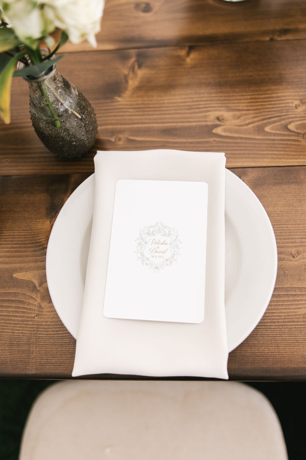 A white plate with a white napkin and a printed menu on top, placed on a wooden table with a dark vase filled with white flowers to the side.