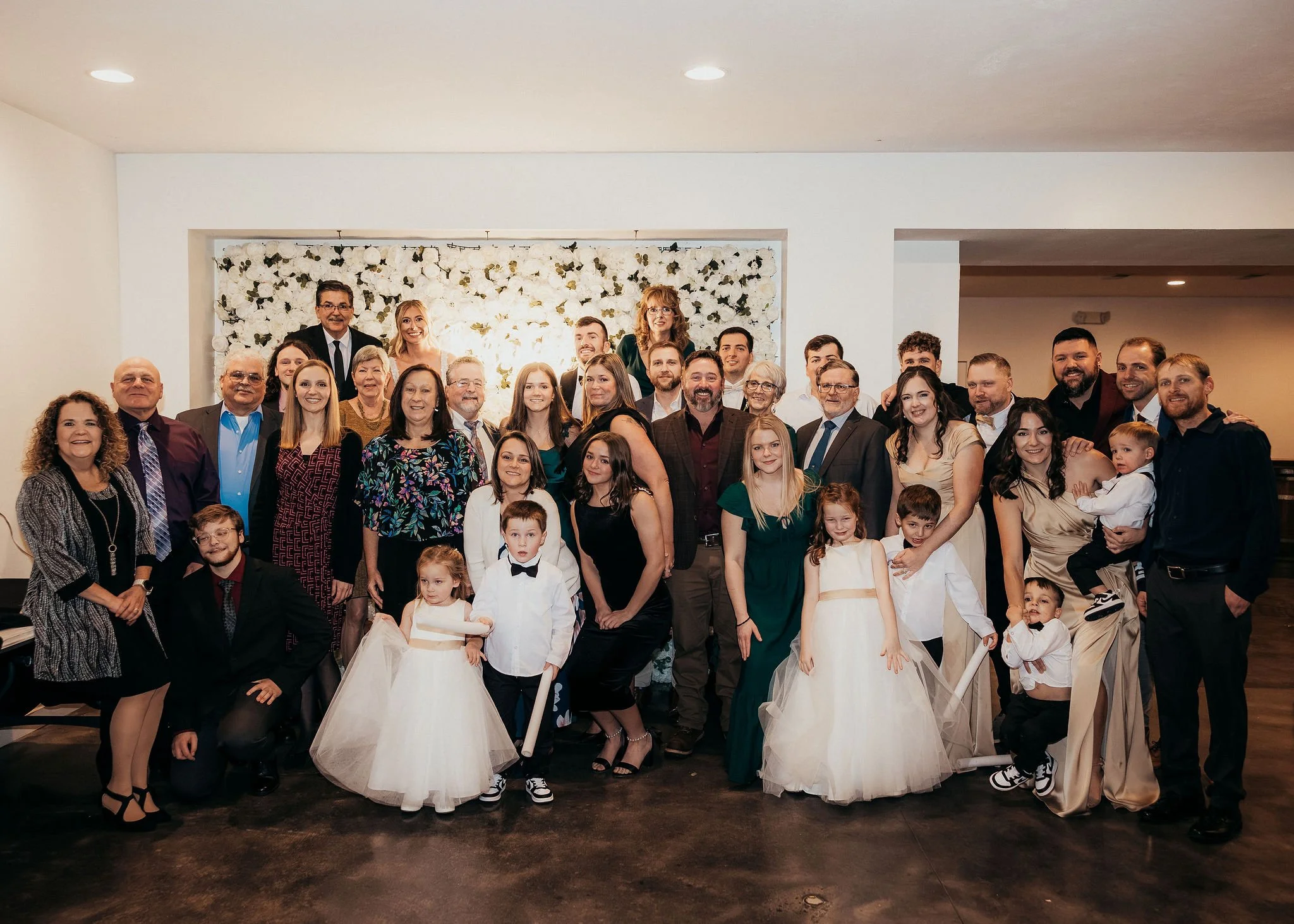 Group of people gathered for a celebration, including adults and children dressed in formal and festive attire, with some children in white dresses, in a decorated indoor space.