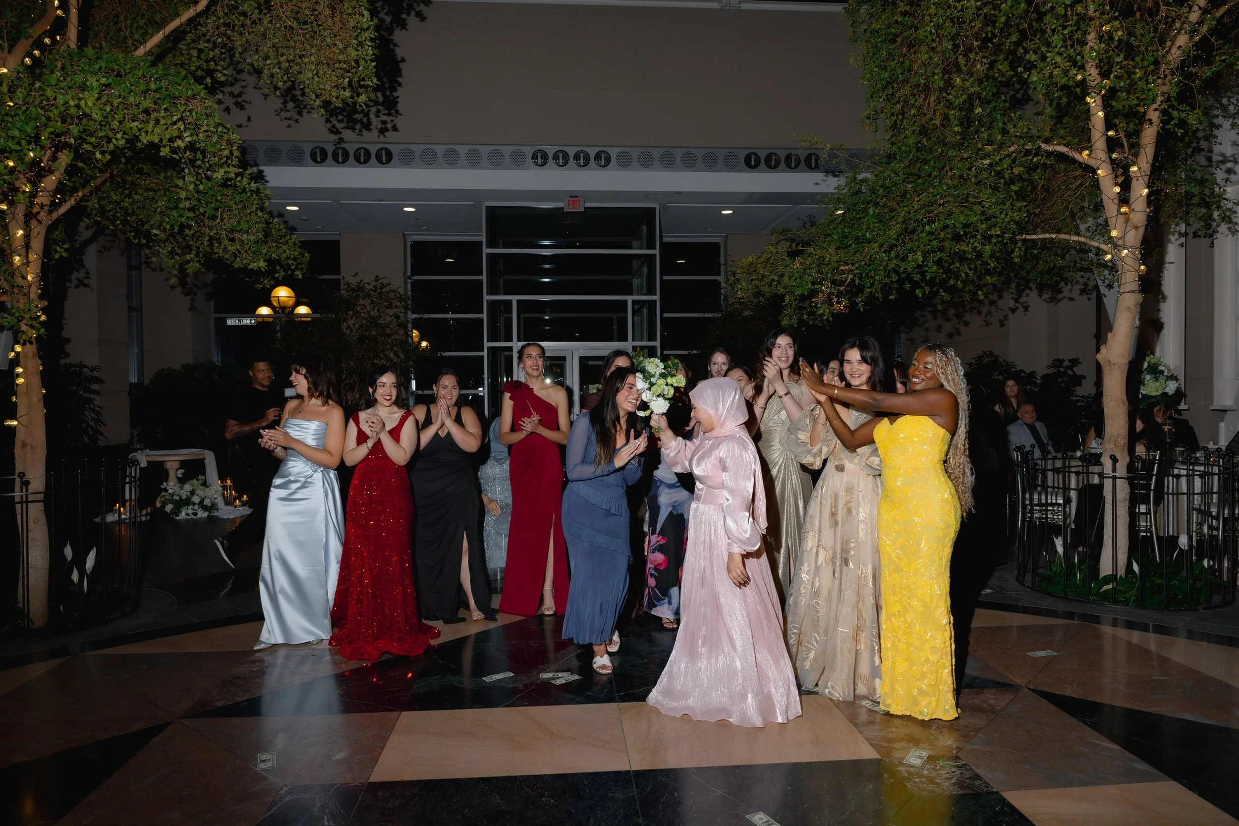 Group of women in elegant dresses celebrating at a wedding reception, with a wedding bouquet, indoor setting, evening lighting, trees with string lights in background.