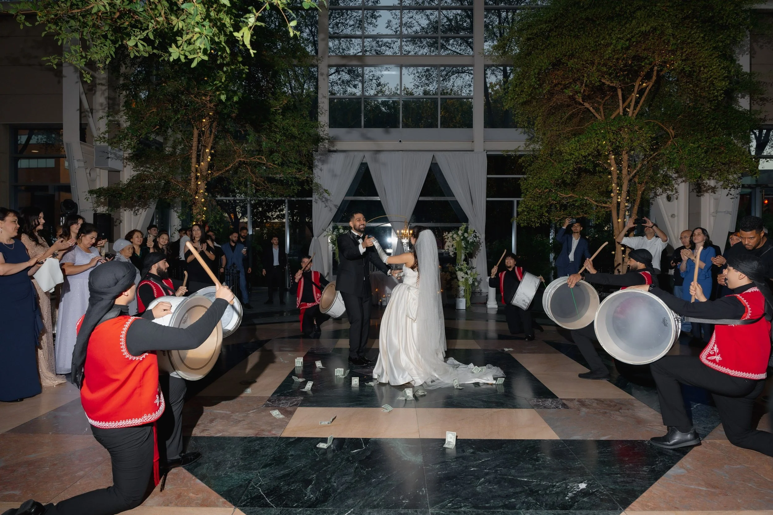 A wedding ceremony taking place indoors with the bride and groom dancing in the center, surrounded by guests watching and taking photos. There are musicians playing drums nearby, and dollar bills are scattered on the floor, indicating a celebratory e