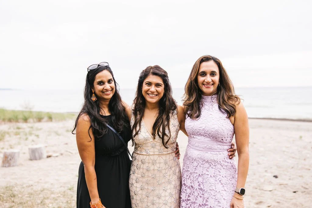 Three women dressed in elegant dresses standing close together on a beach with the ocean in the background, smiling for the camera.