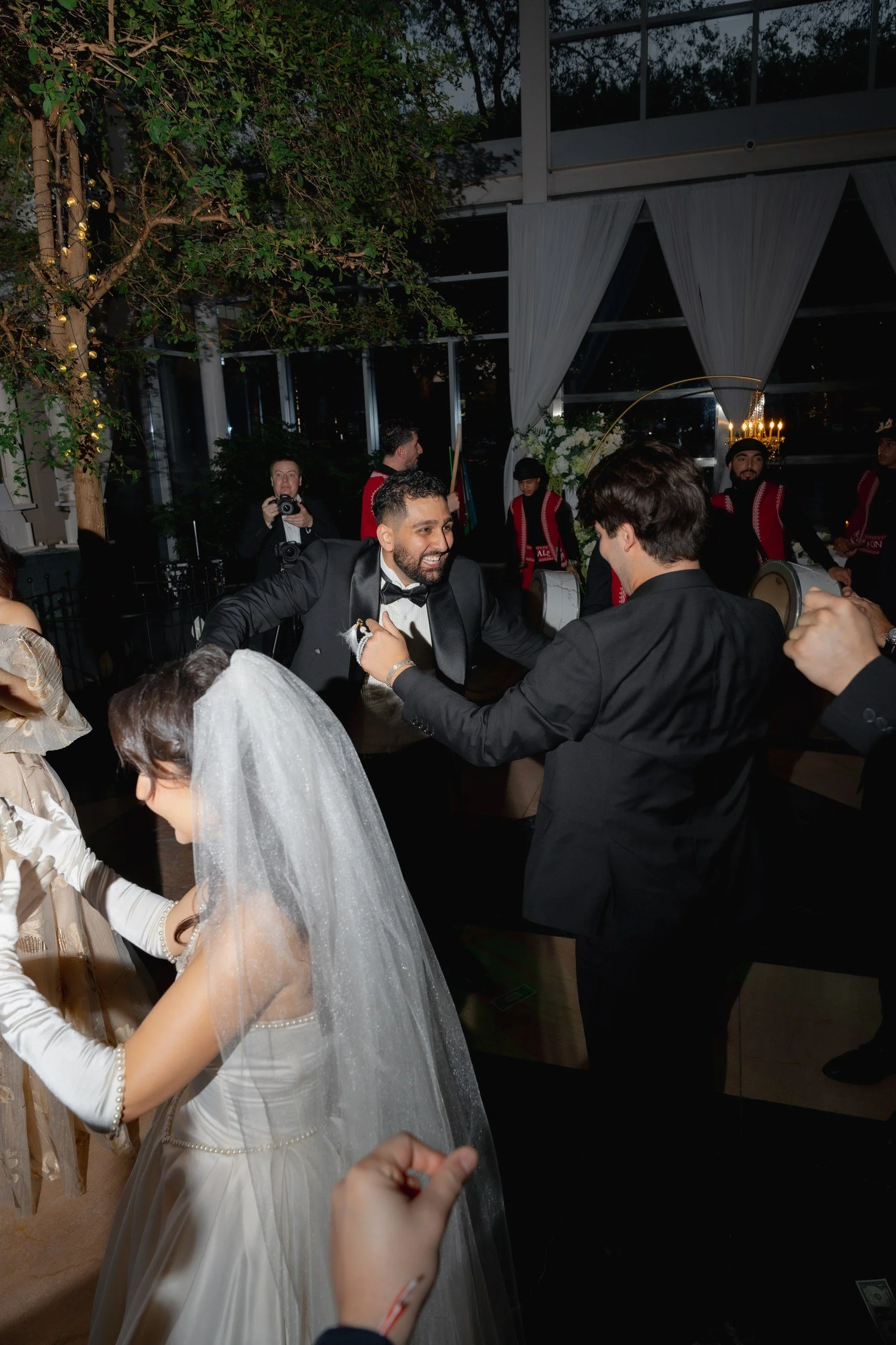 People dancing at a wedding reception, including a man in a tuxedo and a woman in a wedding dress with a veil, with other guests and musicians playing instruments in the background.