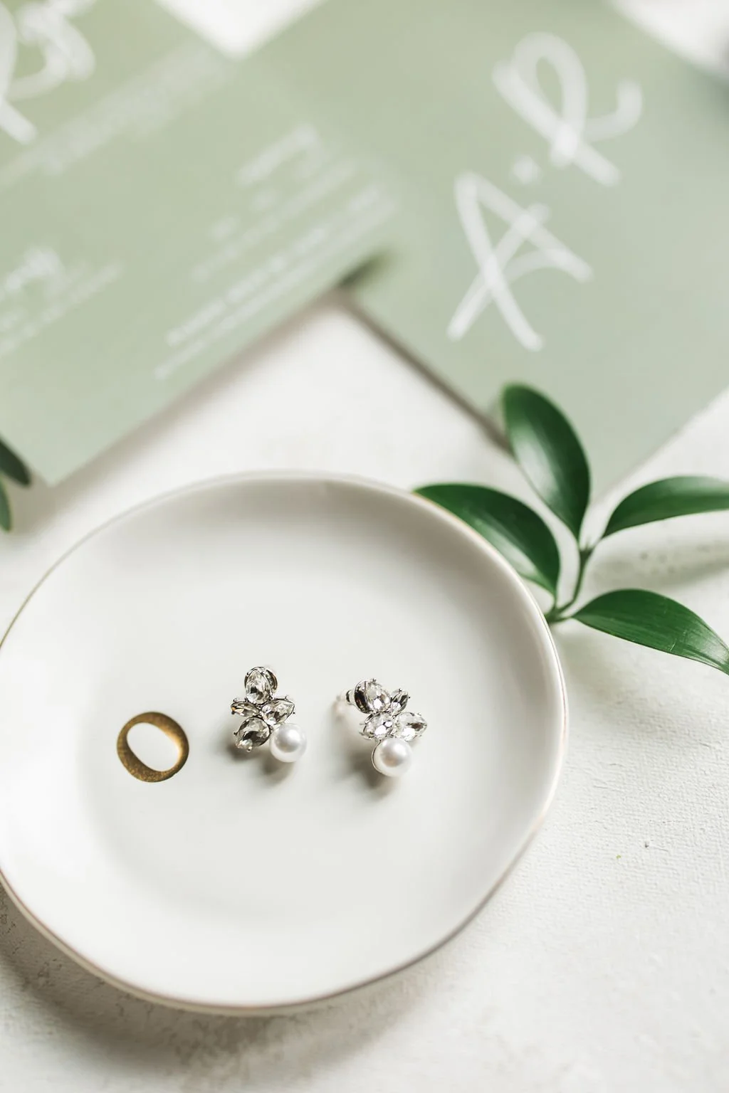 Pair of crystal and pearl earrings on a white dish with green leaves and a blurred green box in the background.