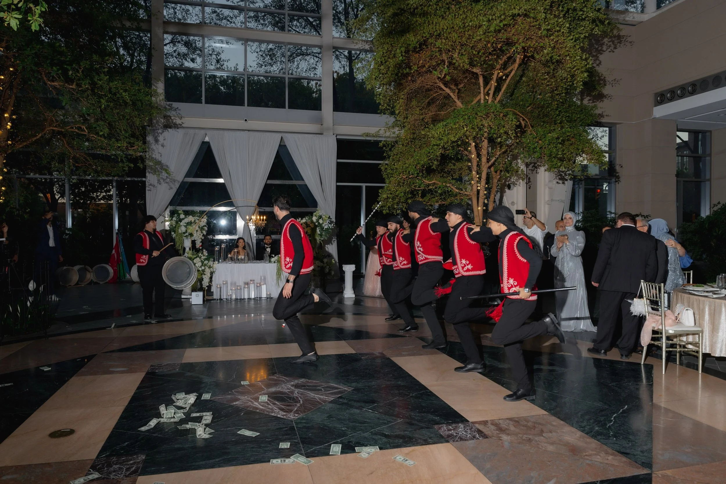 A group of men in traditional red and black costumes performing a cultural dance indoors during an event, with a decorated table, women in elegant dresses, and curtains in the background.