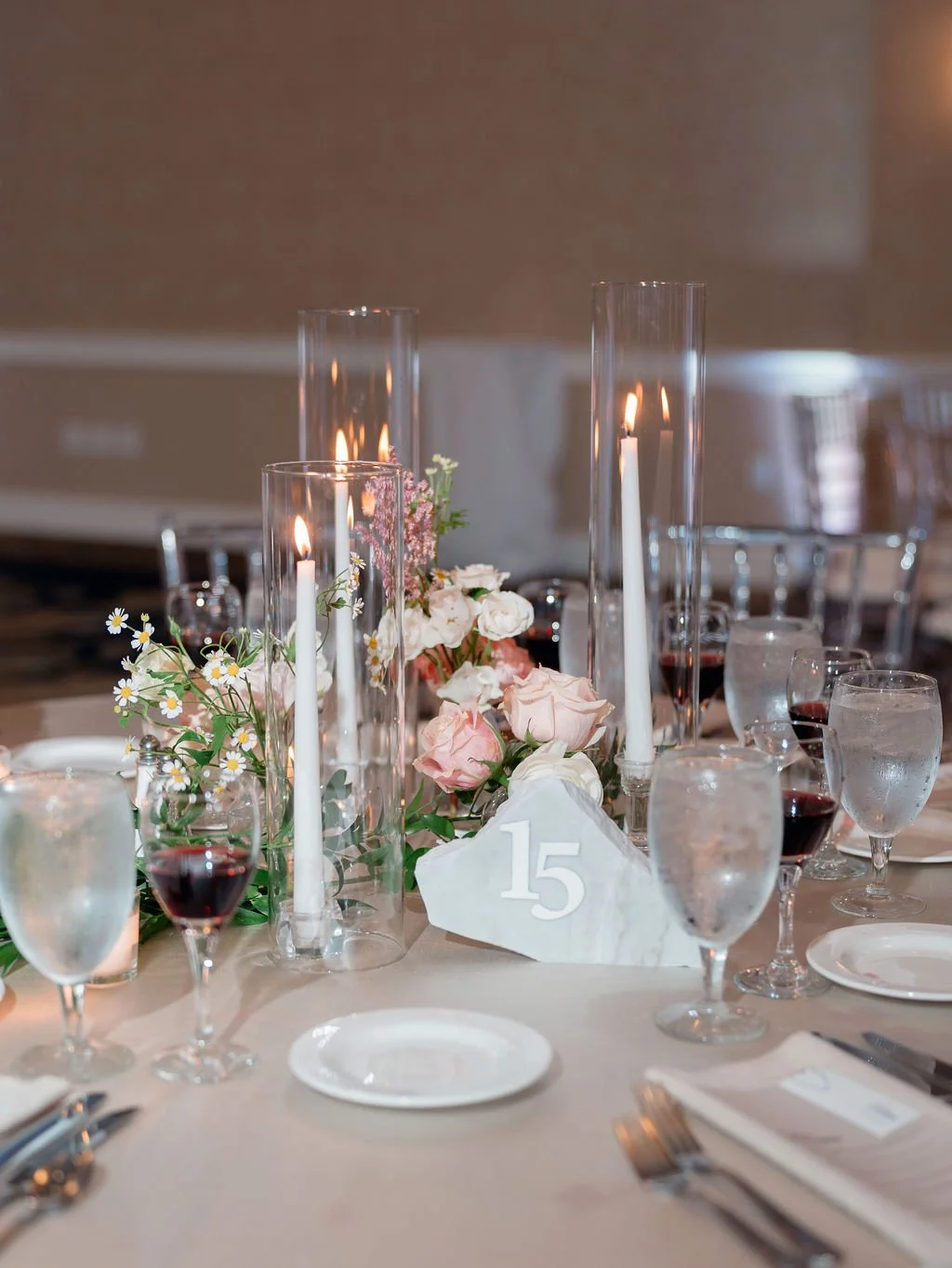 Elegant dinner table decorated with pink and white flowers, tall glass candle holders with white candles, wine glasses filled with red wine, and water glasses, numbered table setting with the number 15.