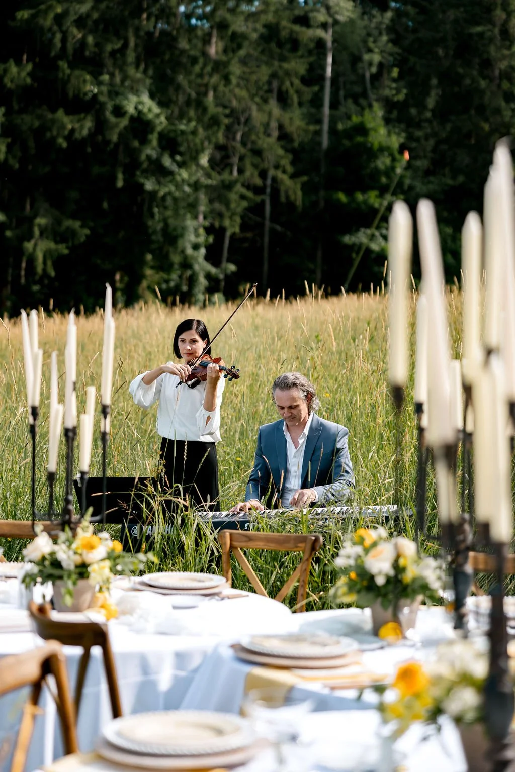 A woman playing the violin and a man playing a keyboard outdoors in a grassy field surrounded by trees, with a table set for a meal in the foreground.