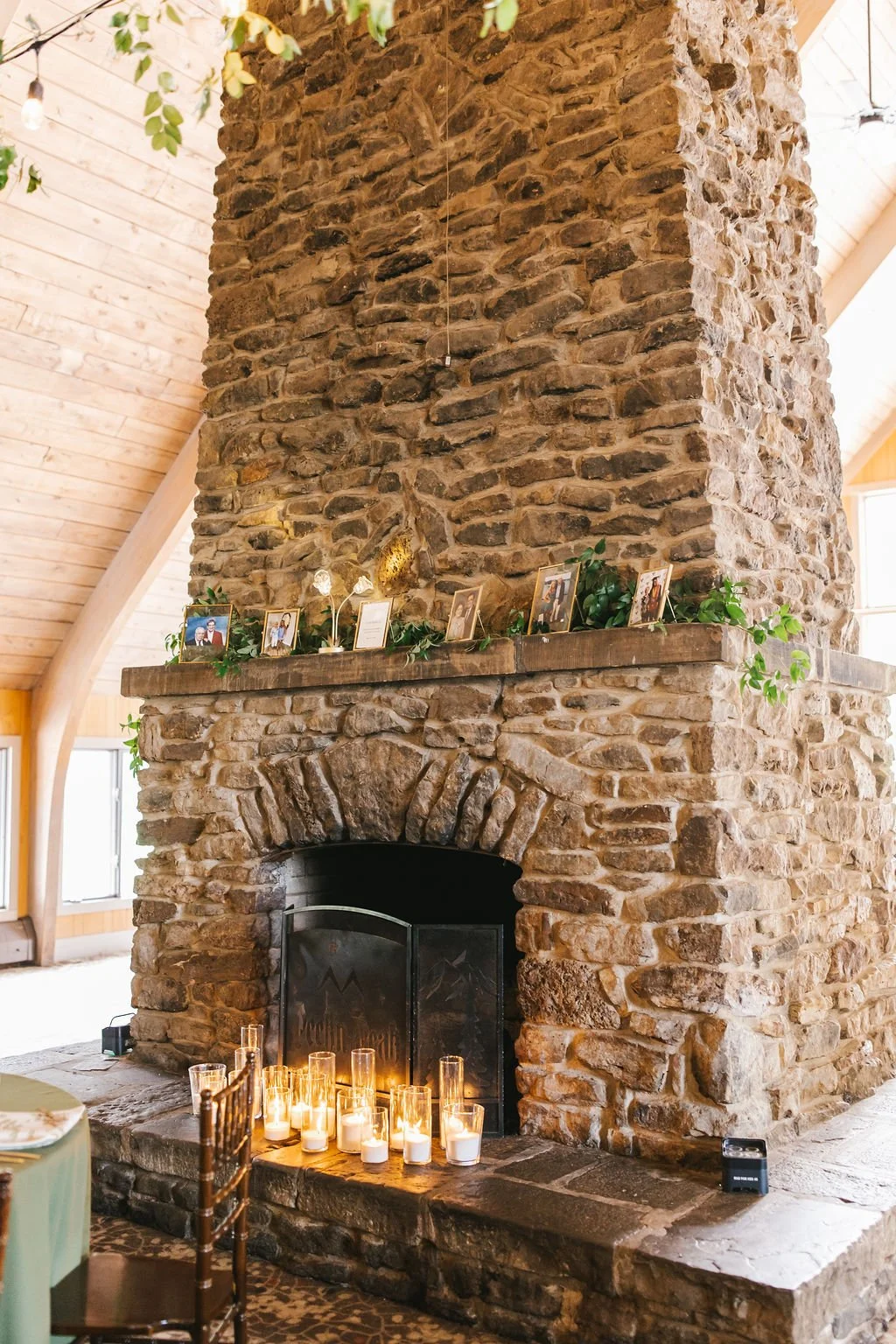 A rustic stone fireplace with a wooden mantel decorated with framed photos and greenery, lit candles on the hearth, and a wooden chair nearby in a cozy room with wood-paneled walls and windows.