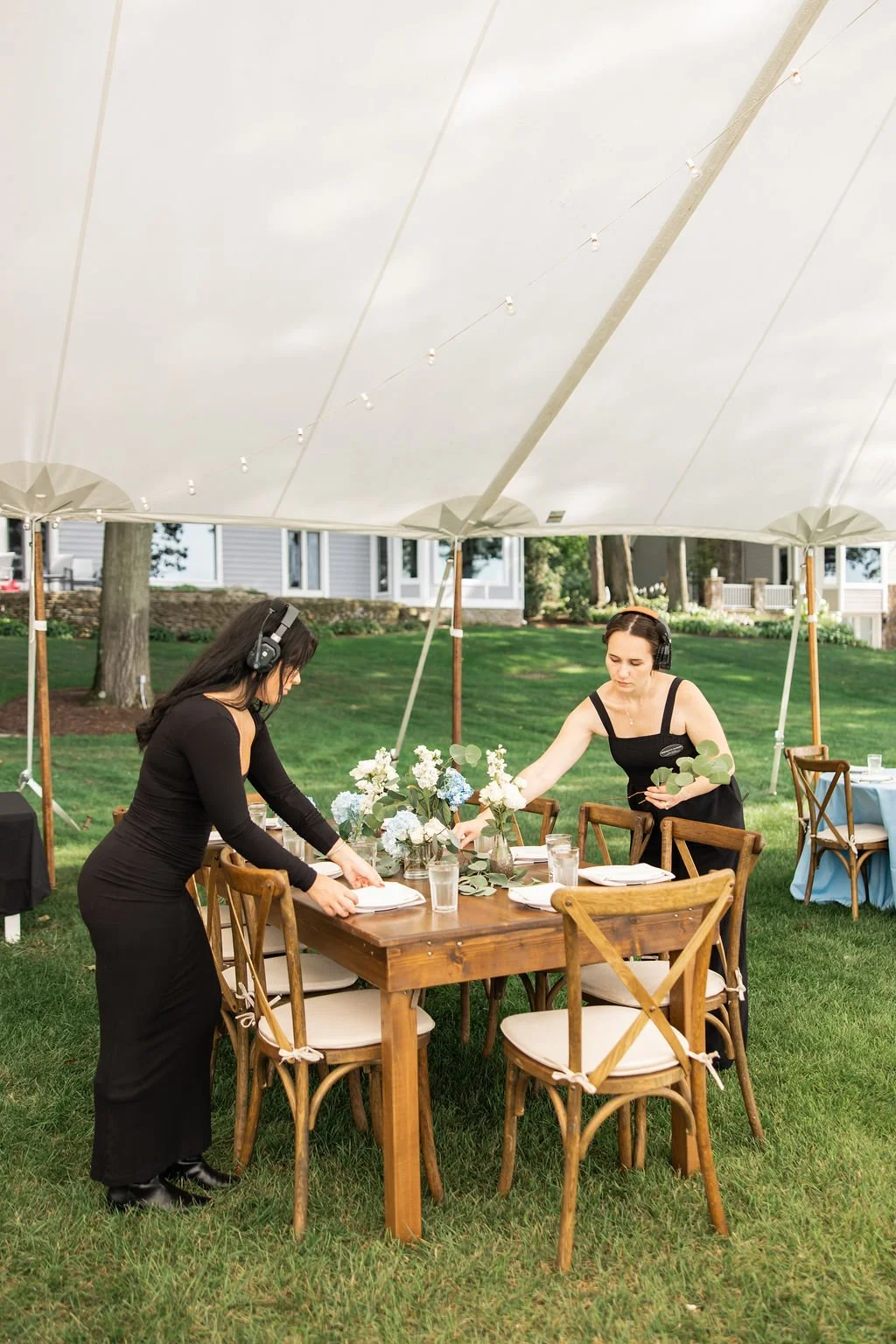 Two women setting up an outdoor event table under a large white canopy or tent, with wooden chairs, floral arrangements, and place settings on a grassy lawn.