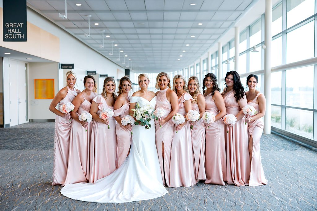 Bride in white wedding dress and nine bridesmaids in pink dresses inside a modern building with large windows.