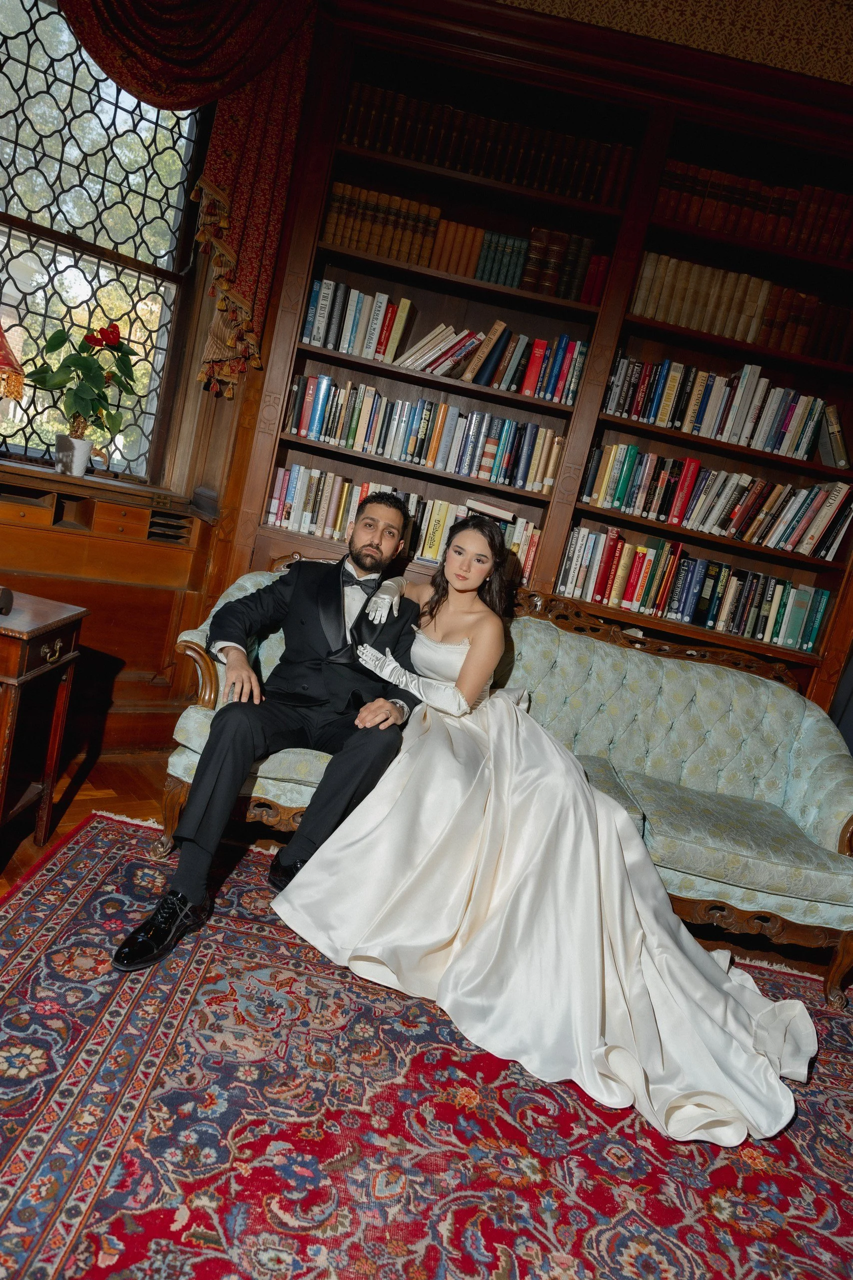 A bride and groom sit together on a vintage sofa in a library, with the groom in a tuxedo and the bride in a white wedding gown, surrounded by a large bookcase filled with books and a Persian rug on the floor.