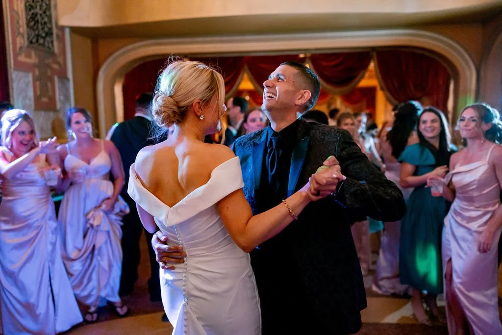 A joyful couple dancing at their wedding reception, surrounded by guests in elegant dresses.