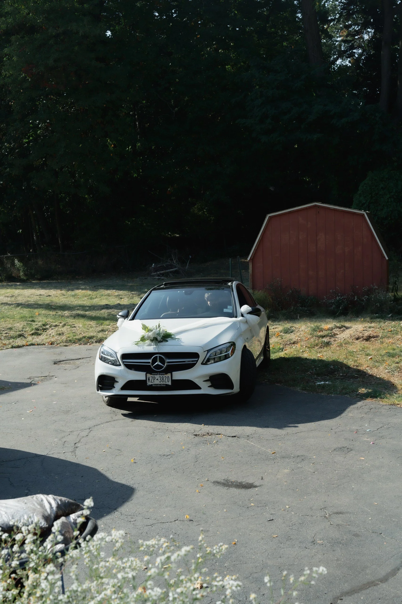 White Mercedes-Benz car parked on a paved driveway with wedding flowers on the hood, in front of a red shed and lush green trees in the background.