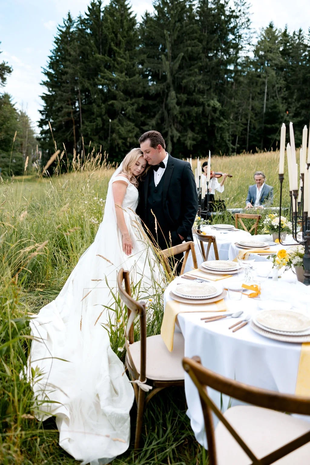 A newlywed couple in wedding attire sharing a tender moment in a field, next to a decorated outdoor dining table with candles, tableware, and floral arrangements, with musicians performing in the background and tall trees surrounding the scene.